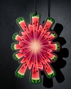 A decorative hanging ornament shaped like a watermelon slice fan, featuring a red and pink watermelon pattern with green rinds and black seeds, suspended against a dark gray background with a strong shadow.