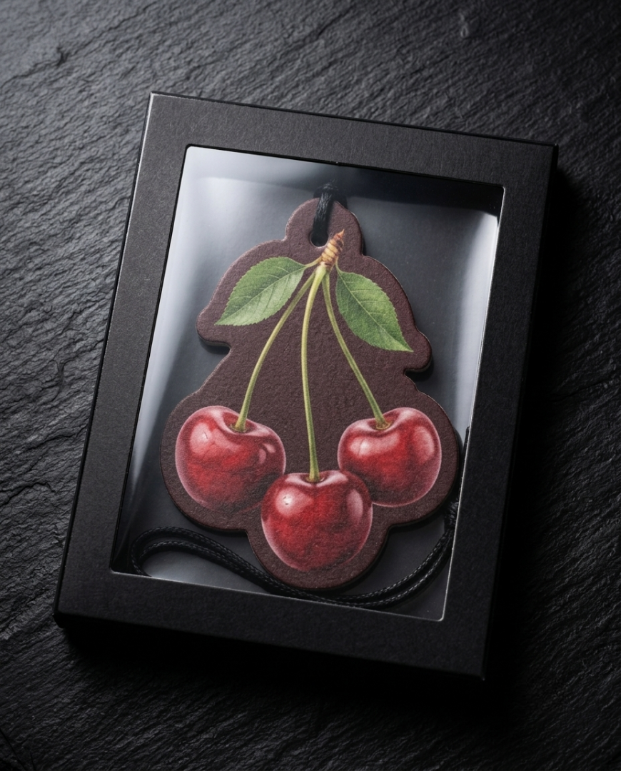 A cherry-shaped leather car air freshener inside a black presentation box with a clear window, set on a dark slate background.