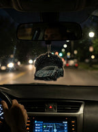 A car air freshener featuring a black sports car with gray and white graphic patterns, hanging from the rearview mirror inside a car at night.
