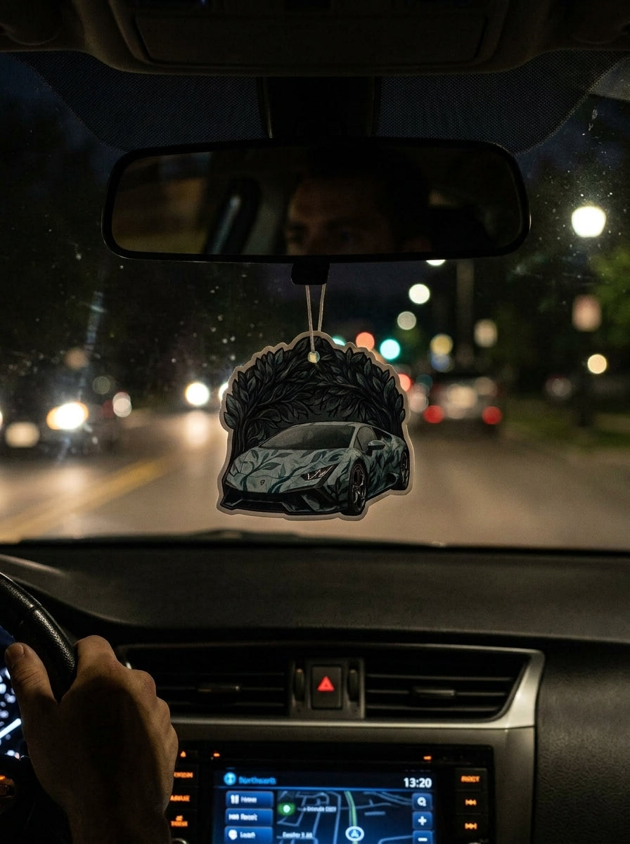 A car air freshener featuring a black sports car with gray and white graphic patterns, hanging from the rearview mirror inside a car at night.