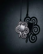 A black metal pendant necklace featuring a stylized four-leaf clover design with swirling patterns and a central rectangular element, shown hanging against a dark textured background with a prominent shadow.