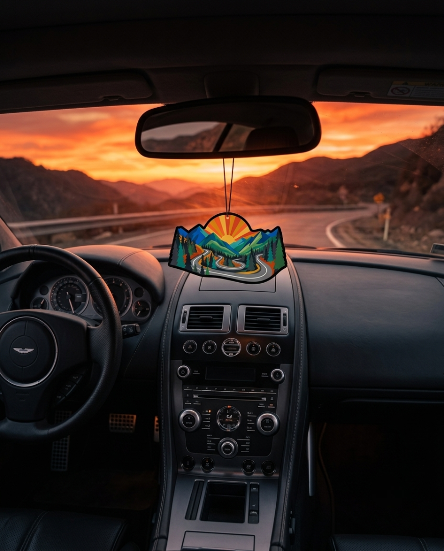 A colorful mountain landscape car air freshener hangs from the rearview mirror inside a car dashboard, with a winding road and mountains visible through the windshield at sunset.