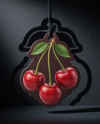 A cherry-shaped hanging ornament or car air freshener with three glossy red cherries, green leaves, and stems, suspended by a black cord against a dark gray background with a shadow.