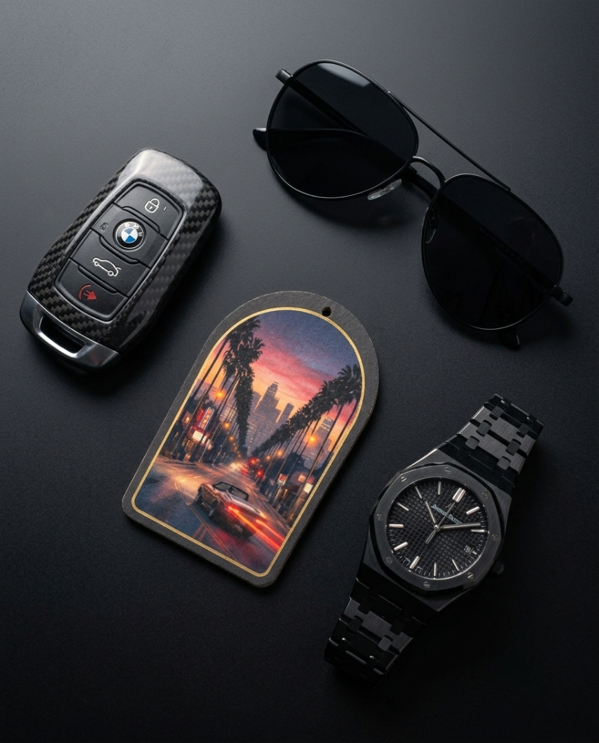 A styled flat-lay of luxury accessories on a dark background, including a black carbon-fiber-look car key fob with a BMW roundel, black aviator sunglasses, a rectangular car license plate frame featuring a sunset city street scene, and a black watch with a matching black digital bracelet.