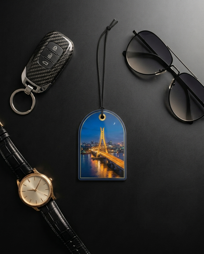 A car key fob, sunglasses, a wristwatch, and a car air freshener tag are arranged on a dark gray background. The air freshener tag features a printed image of a illuminated bridge at night over water.