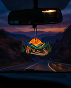 A car air freshener hanging from the rearview mirror, featuring a scenic mountain landscape with a winding road and a bright orange sunburst, set against a blurred mountain road at dusk.
