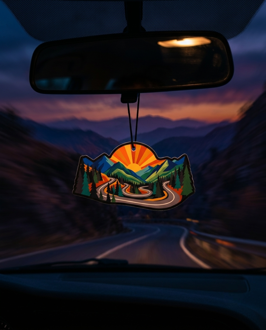 A car air freshener hanging from the rearview mirror, featuring a scenic mountain landscape with a winding road and a bright orange sunburst, set against a blurred mountain road at dusk.
