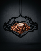 A copper-colored Celtic knot pendant with a central cup holder, suspended by a black cord against a dark gray background with a dramatic shadow.