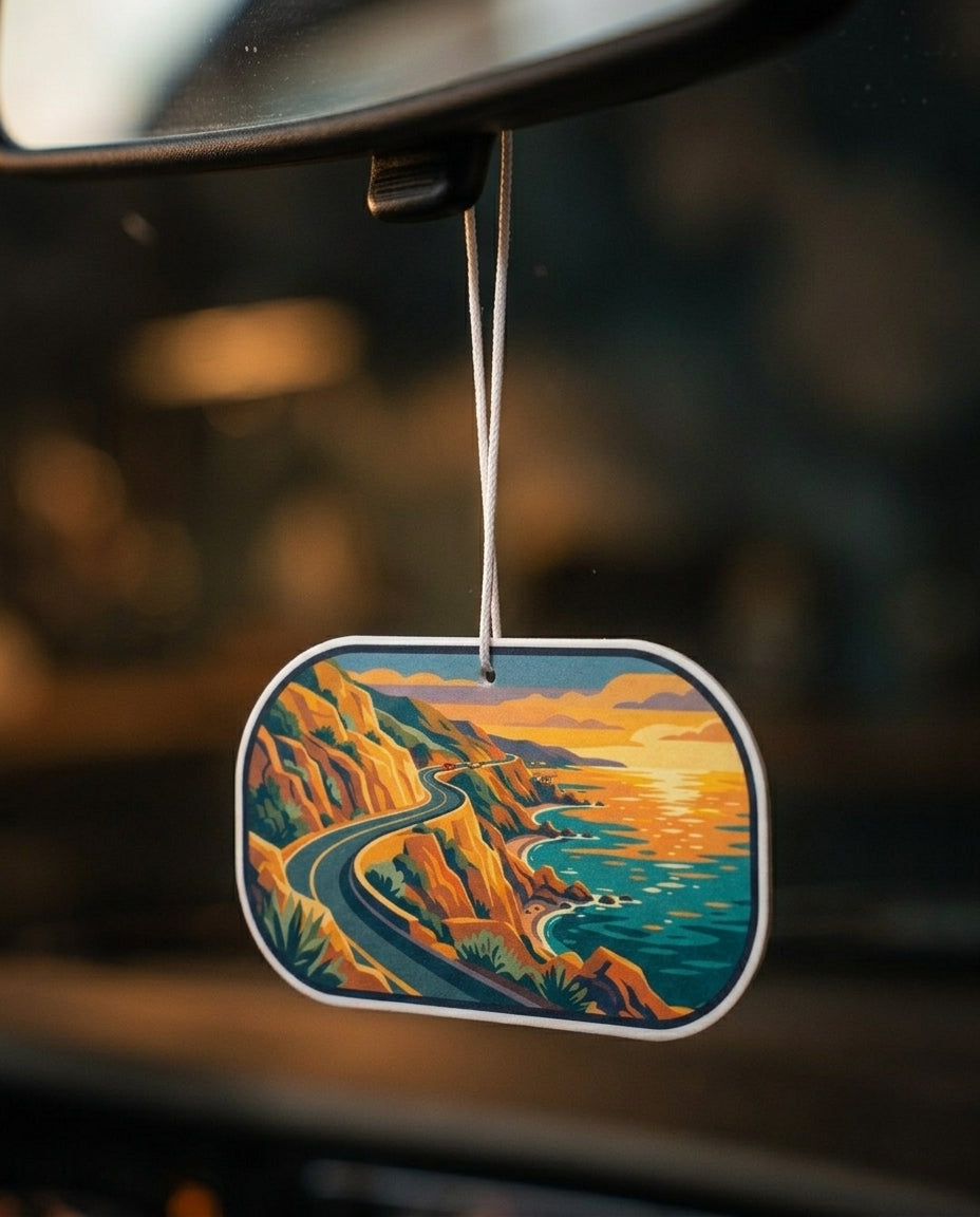 A car air freshener hanging from a rearview mirror. The oval-shaped fragrance tag features a stylized illustration of a winding road along a coastal cliffside at sunset, with warm orange and yellow tones on the cliffs and sky, and deep teal-blue tones in the water and lower landscape.