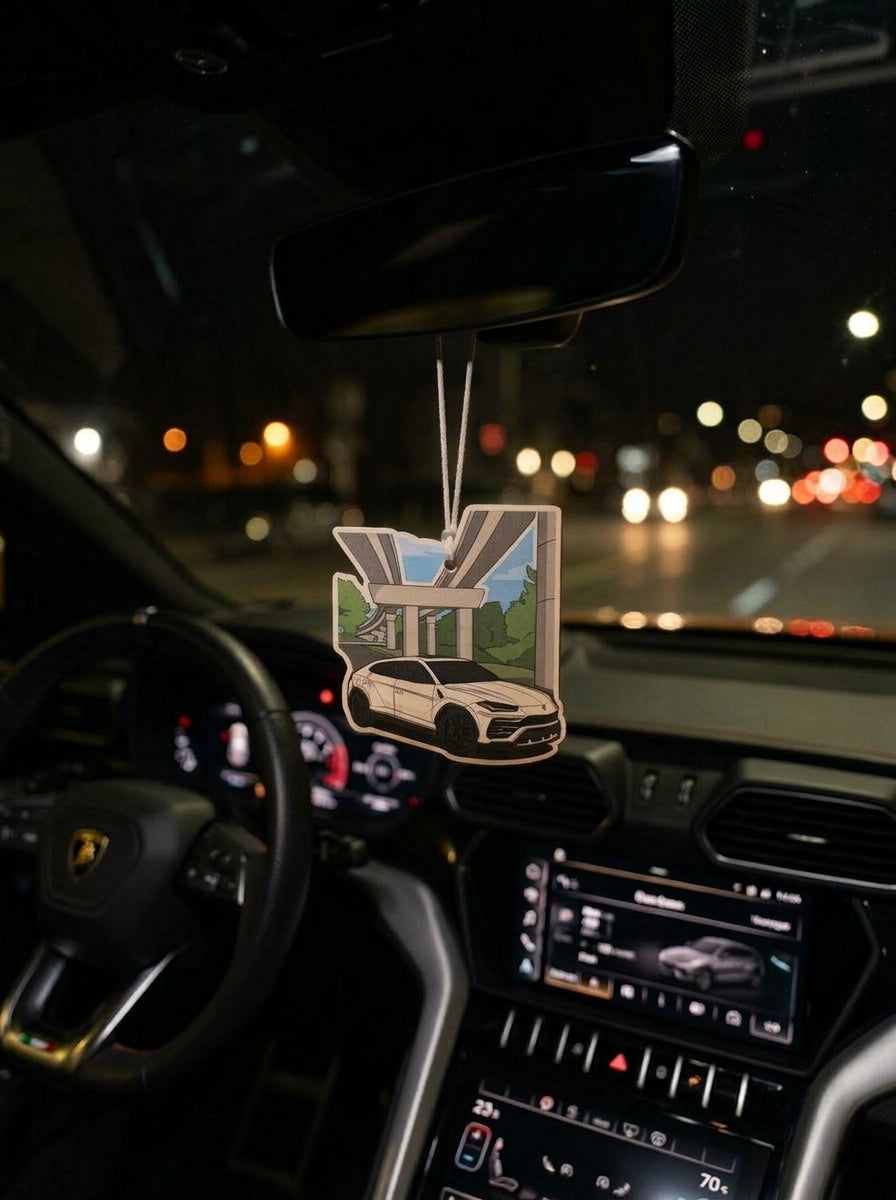 A car air freshener featuring a white sports car under a bridge with green trees, hanging from the rearview mirror inside a car at night with blurred street lights outside.