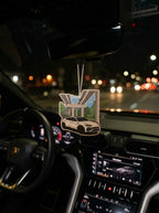 A car air freshener featuring a white sports car under a bridge with green trees, hanging from the rearview mirror inside a car at night with blurred street lights outside.