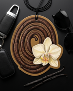 A round wooden car air freshener ornament with a hand-painted brown wood grain swirl pattern and a white orchid flower, styled on a black background with sunglasses, a keychain, and vanilla beans.