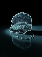 A die-cut car-shaped sticker or patch featuring a stylized sports car under a tree canopy, shown on a glossy black reflective surface with a cord attached.