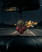 A cherry-shaped car air freshener hangs from a rearview mirror inside a moving car, with blurred city lights and a highway road visible outside at night.