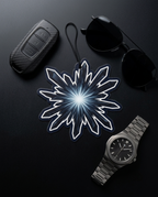 A styled flat-lay featuring a central white and blue glowing snowflake-shaped air freshener or ornament, surrounded by a black carbon-fiber car key fob, black sunglasses, and a silver metal wristwatch on a dark background.