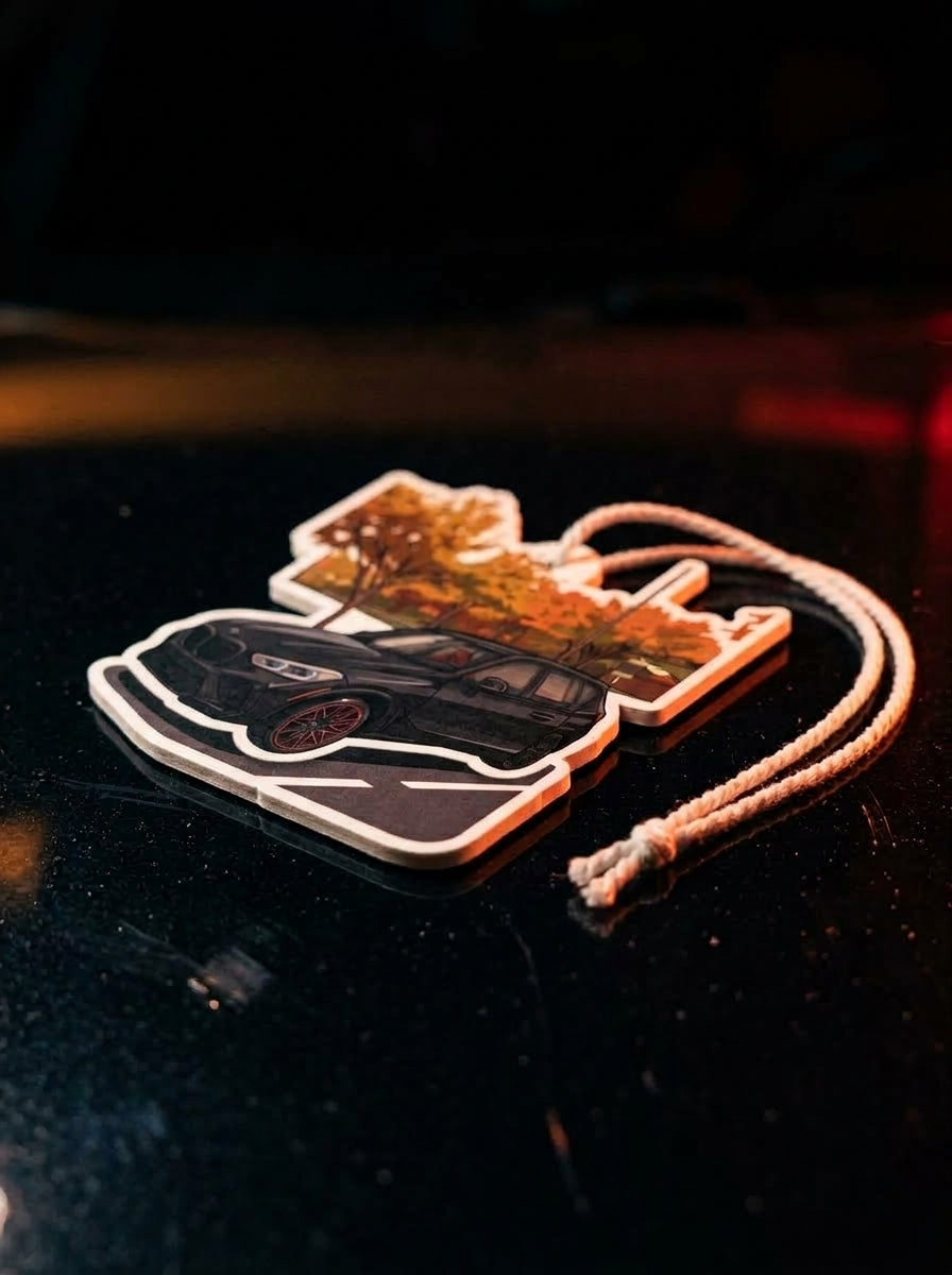 A die-cut car-shaped air freshener or ornament featuring a black car and autumn tree design, with a white cord, resting on a dark reflective surface.