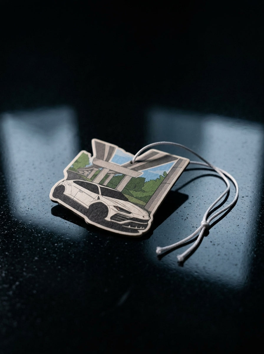 A small rectangular car-shaped air freshener or charm with a printed white sports car and green landscape scene, attached to a thin cord, photographed on a dark reflective surface.