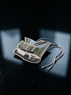 A small rectangular car-shaped air freshener or charm with a printed white sports car and green landscape scene, attached to a thin cord, photographed on a dark reflective surface.
