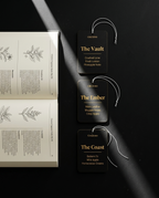 Three black fragrance tag cards with gold text for Creations Cinnamon, The Vault, and The Ember, displayed next to an open book on a dark background with dramatic diagonal light beams. A third tag card is labeled 'The Coast'.