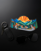 A colorful mountain landscape patch with a winding road, trees, and a rising sun, styled on a dark background with a black leather key fob and keys, and black sunglasses.