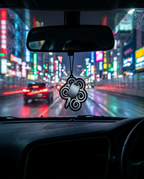A black and white decorative car air freshener hanging from the rearview mirror inside a car, with a blurred city street at night featuring neon lights in the background.