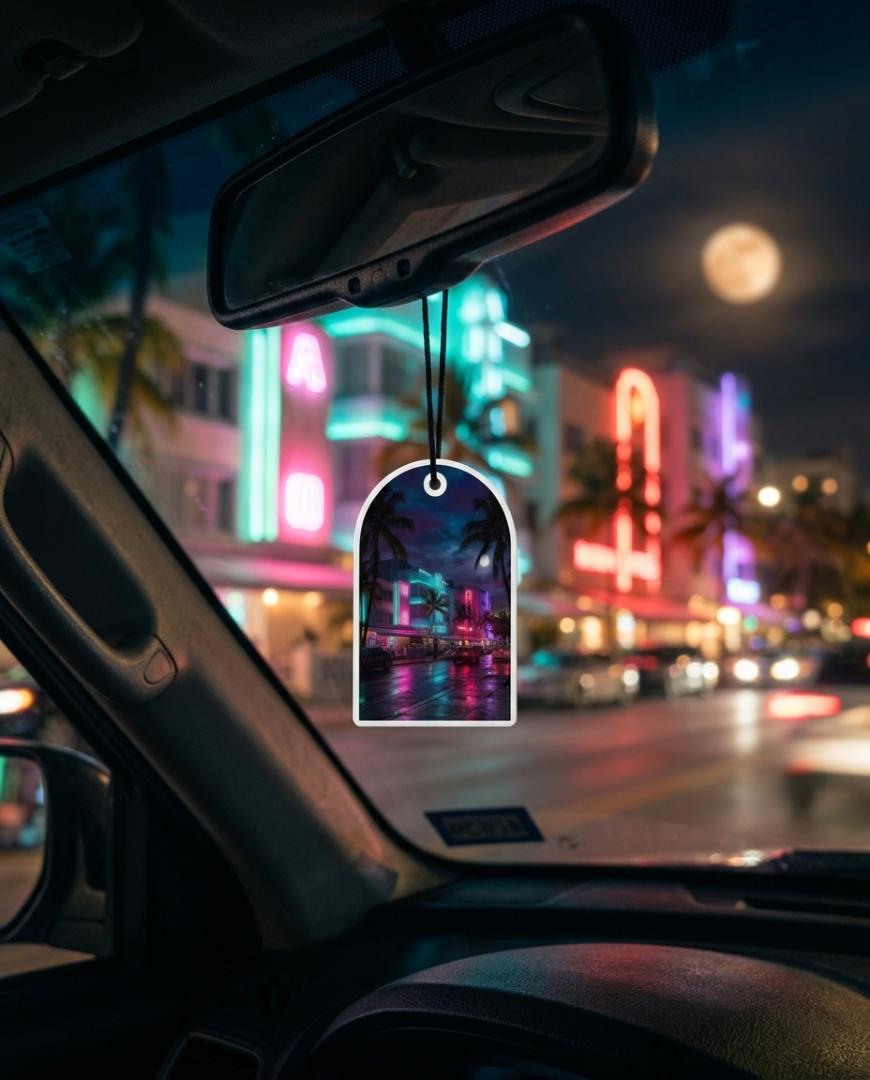 A neon-themed car air freshener hanging from a rearview mirror in a car, with a blurred Miami-style night street scene outside.