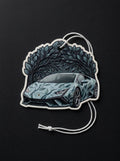 A die-cut car air freshener or ornament featuring a stylized Lamborghini-style sports car with a blue and gray leaf pattern, set against a dark leafy illustration. It has a white hanging cord and is photographed on a dark textured background.
