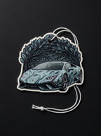 A die-cut car air freshener or ornament featuring a stylized Lamborghini-style sports car with a blue and gray leaf pattern, set against a dark leafy illustration. It has a white hanging cord and is photographed on a dark textured background.