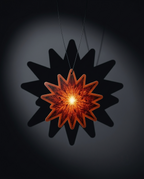 A glowing star-shaped pendant or ornament with an amber-orange translucent center and a dark outer border, suspended against a dark gray background with a large black star shadow behind it.