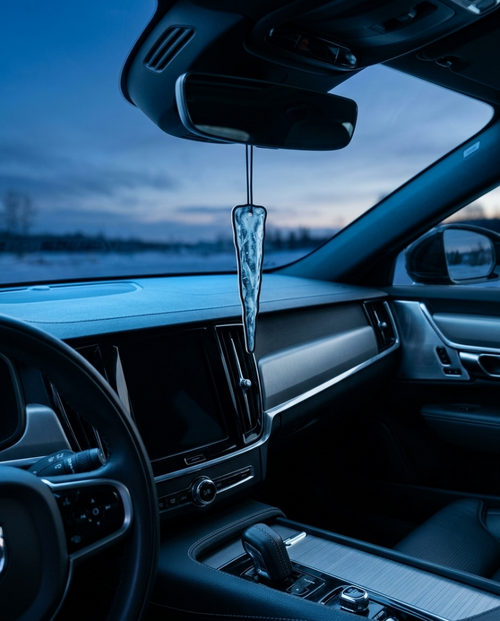 A clear, textured car air freshener hangs from the rearview mirror inside a modern car cabin at dusk.