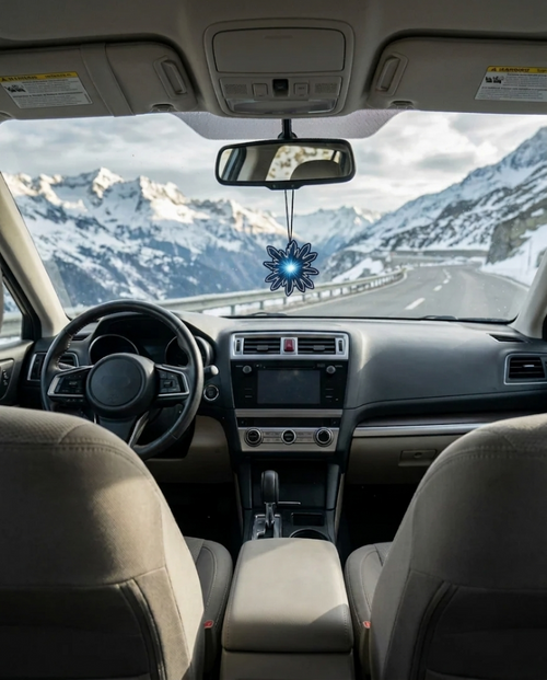 A snowflake-shaped blue air freshener hanging from the rearview mirror inside a car, with a snowy mountain road visible through the windshield.