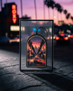 A rectangular clear acrylic shadow box with a black frame displays a colorful sunset city street scene framed by palm trees. The shadow box sits on a dark textured surface with a blurred city street at dusk in the background.