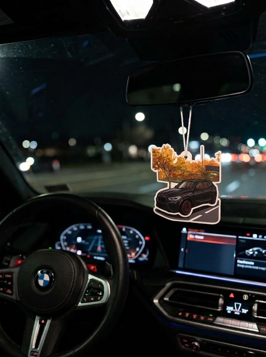 A car air freshener featuring a black SUV with autumn trees, hanging from the rearview mirror inside a BMW car at night.