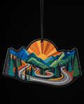 A die-cut mountain landscape air freshener or ornament featuring a winding road through pine trees and hills under a sunburst sky, shown against a black background.
