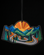 A die-cut mountain landscape air freshener or ornament featuring a winding road through pine trees and hills under a sunburst sky, shown against a black background.