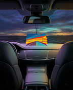 A colorful air freshener or car fragrance diffuser hanging in the center of a car dashboard, with a sunset road scene visible through the windshield.