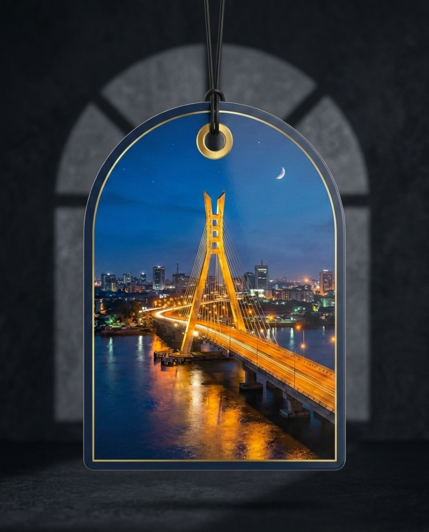 An arched rectangular hanging ornament or tag with a black cord and gold-toned top ring, printed with a nighttime city bridge scene over water. The ornament is centered against a dark gray arched backdrop.