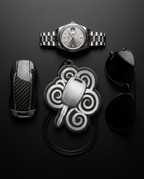 A styled flat-lay of luxury accessories on a dark background, including a silver Rolex-style wristwatch, a black carbon-fiber-patterned case, a silver key fob in a decorative cloud-shaped holder, and black sunglasses.