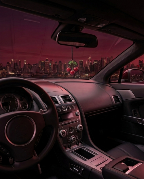A dark car interior with a cherry-shaped air freshener hanging from the rearview mirror, set against a pink-tinted city skyline at night visible through the windshield.