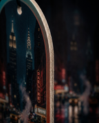 A close-up of an arched decorative tag or ornament featuring a New York City skyline print with illuminated skyscrapers and a Daellire theater sign, shown against a blurred dark city background.
