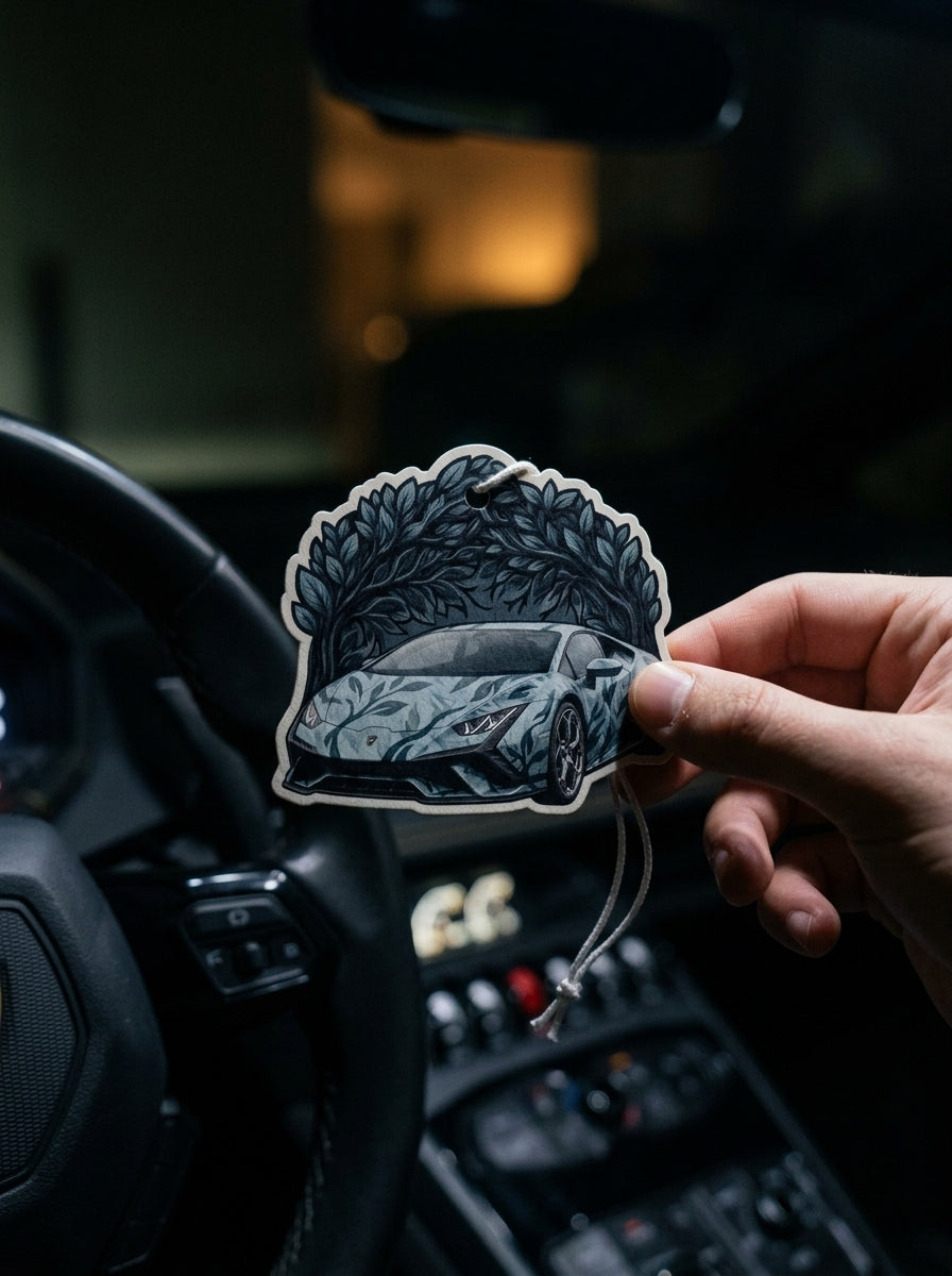 A hand holds a die-cut car air freshener sticker featuring a stylized gray sports car under a leafy arch, shown in front of a dark car interior with a steering wheel and dashboard.