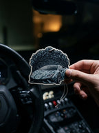 A hand holds a die-cut car air freshener sticker featuring a stylized gray sports car under a leafy arch, shown in front of a dark car interior with a steering wheel and dashboard.