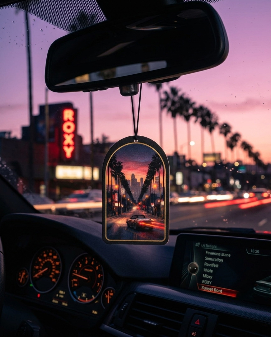 A car air freshener hanging from the rearview mirror in a car during sunset. The clear acrylic air freshener features a printed scene of a city street lined with palm trees at dusk, with a car driving down the road.