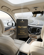 A car interior with a hanging air freshener tag reading 'The Coast' and scent notes 'Balsam Fir', 'Wild Apple', and 'Herbaceous Greens'. The tag is suspended in front of the windshield, with cream-colored seats and a coastal road visible outside.