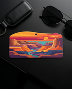 A colorful landscape-themed keychain or card holder with a canyon scene, displayed on a dark surface alongside sunglasses, a black leather key fob, and a phone.