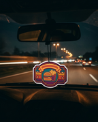 A colorful burger-themed car air freshener hangs from the rearview mirror on a car dashboard during a night highway drive.