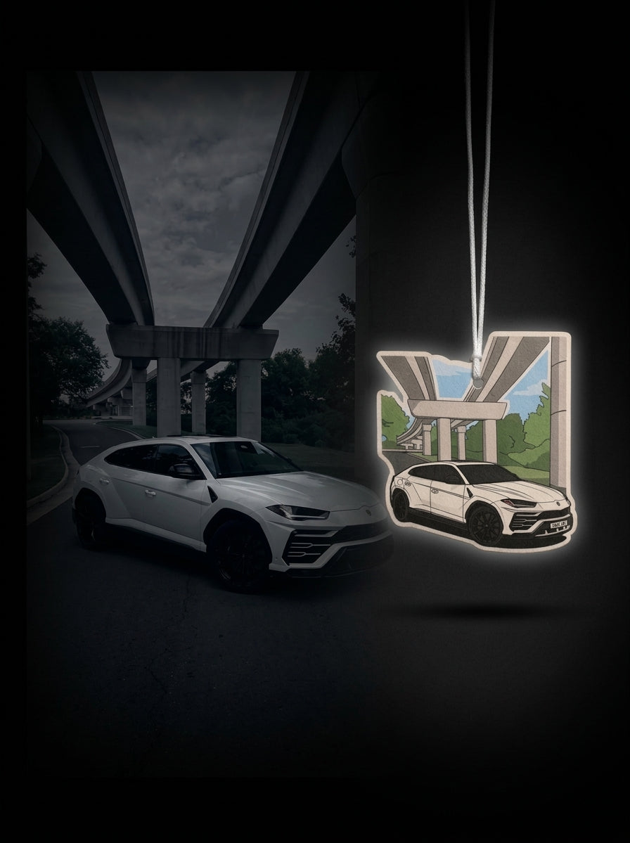 A car-shaped hanging ornament featuring a white Lamborghini Urus with a printed scene of a white car under an elevated highway overpass, shown next to a real white Lamborghini Urus in a dark outdoor setting.