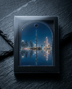 A square black-framed art print or picture frame displaying a nighttime city skyline with a tall central tower and illuminated buildings reflected in water, set against a dark textured stone background.
