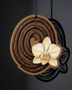 A decorative oval wall hanging or plaque with a dark brown wood-grain spiral design and a cream-colored orchid flower attached near the lower right, shown hanging against a dark gray background.