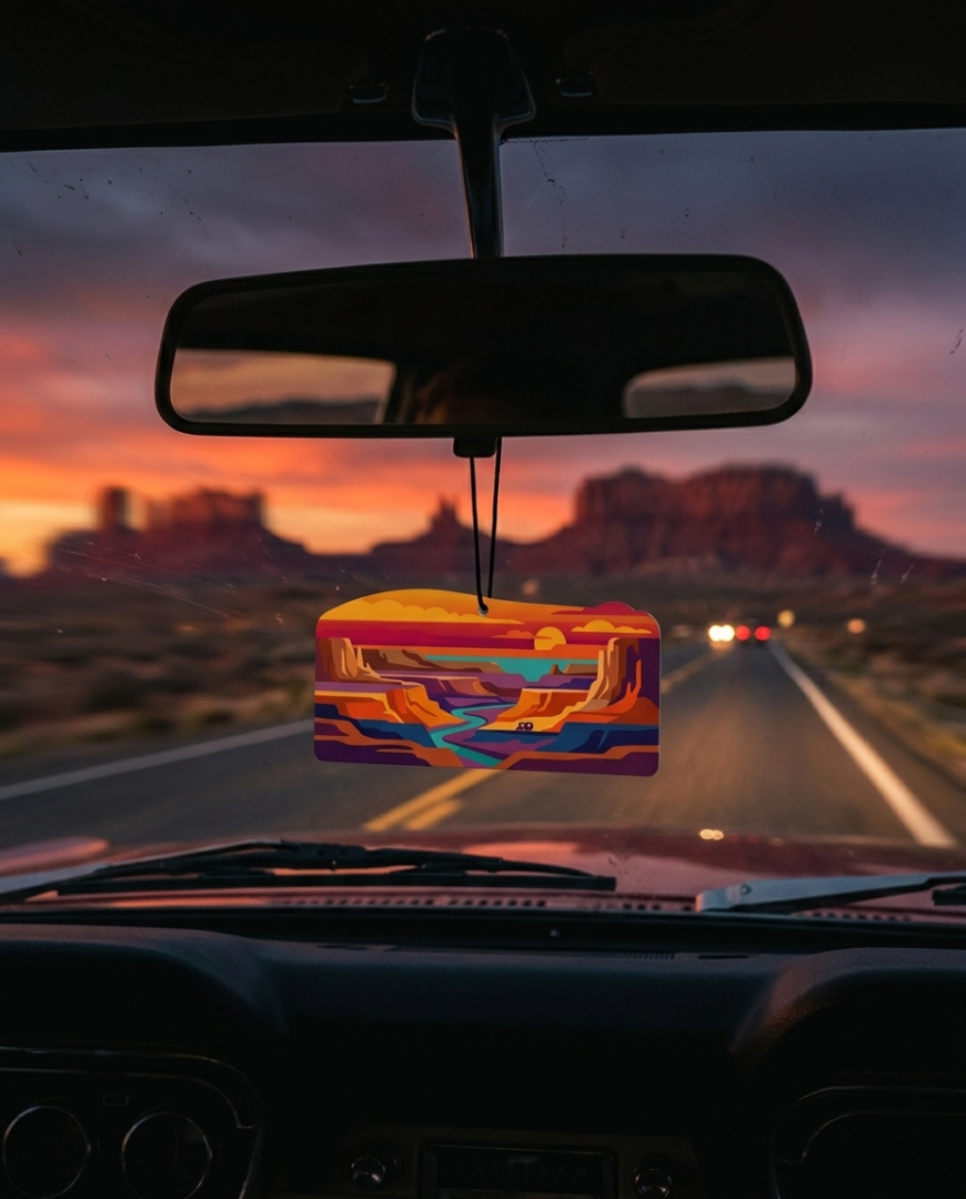 A colorful, geometric-patterned car air freshener hangs from the rearview mirror on a highway at sunset, with desert rock formations visible in the background.